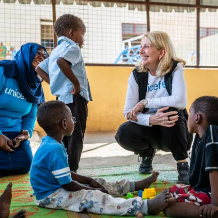 A woman in a UNICEF shirt smiles while kneeling in front of children in a play area. The warm atmosphere is supportive and engaging.