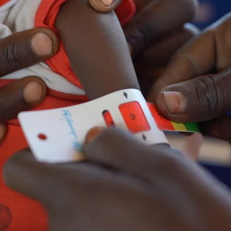 A child's arm is measured with a MUAC tape by two adults, wearing a red and white garment. The scene conveys care and urgency in a healthcare context.
