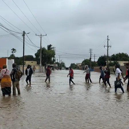 People walking through a flooded street under gray skies. The water covers the road, and they hold hands, conveying a sense of solidarity and resilience.
