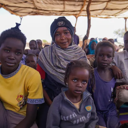 A group of children sit closely together under a makeshift tent. The scene conveys resilience and community.