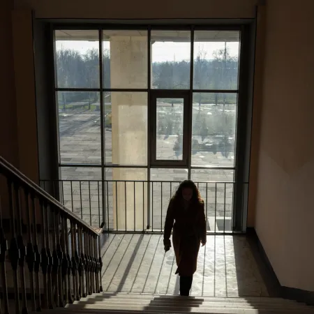 A person walks up a dimly lit staircase, silhouetted against a large window that reveals an overcast outdoor scene. The mood is quiet and contemplative.