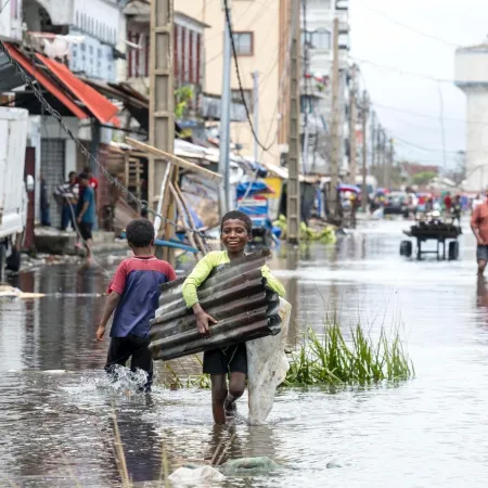 Two children navigate a flooded street, one carrying corrugated metal, both appearing determined. The scene is bustling with people amid submerged cars and buildings.