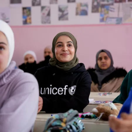 In a classroom with pink walls, a smiling girl wearing a hijab and a UNICEF shirt sits among attentive peers. The atmosphere is warm and positive.