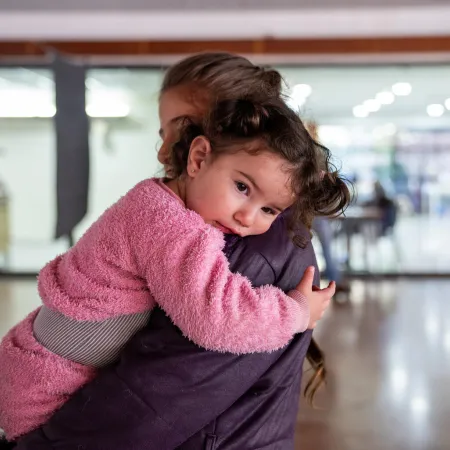 A mother holds a toddler wearing pink clothes in her arms in a brightly lit room. 