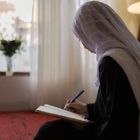 A girl in a white hijab is writing in a notebook, seated on a carpet. 