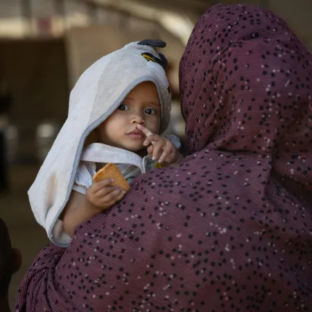 A child with a towel hood is held by a person in a patterned cloak, gazing intently and holding a piece of bread. 