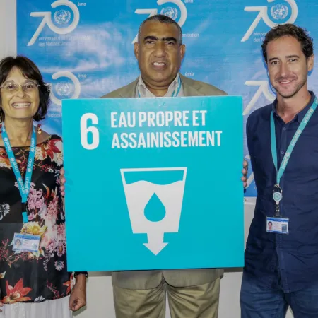 Three people smiling, standing in front of a blue banner, holding a sign that reads "Eau Propre et Assainissement" with the SDG 6 sign that has a water symbol.