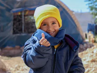 In Syria, 4-year-old Rashid smiles and gives a thumbs-up sign, while wearing his new winter clothes distributed by UNICEF in Fafin camp for displaced families.