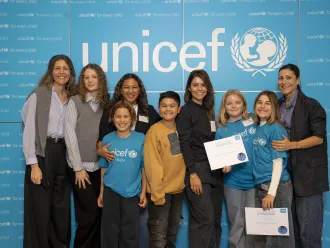 A group of four women and five children smile against a blue UNICEF backdrop. Two kids hold certificates. The scene is joyful and celebratory.