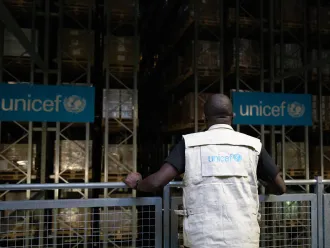 A man stands at a railing with his back to the camera, overlooking a warehouse staked with boxes. He is wearing a vest that says UNICEF.