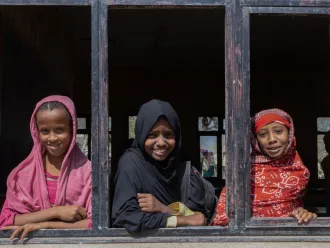 Three young girls with colourful head scarfs on, look out of a wooden window frame - all are smiling.  