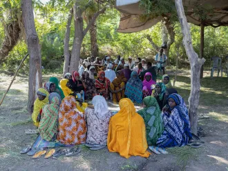 A group of women in colourful clothing sit under the shades of trees and a makeshift shade cover. 