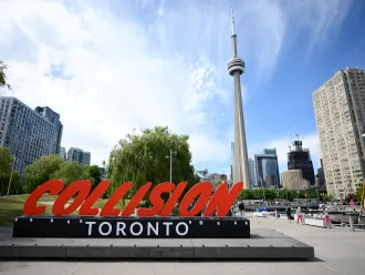 A red and black sign in the foreground reads 'Collision Toronto' in the background the CN tower and some buildings can be seen.  