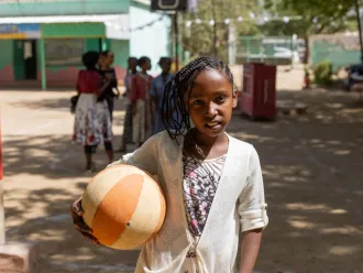 A child stands holding a basketball while smiling at the camera in an outdoor setting with other children standing in the background