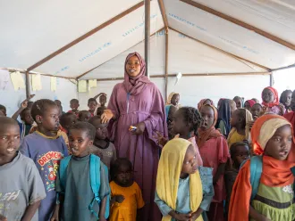 A teacher in a mauve robe stands among young children in a classroom within a UNICEF tent. Children wear colorful clothing.