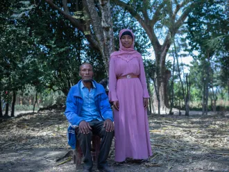 A man sits on a stool and his teenage daughter stands next to him. They are outdoors with many trees behind them.