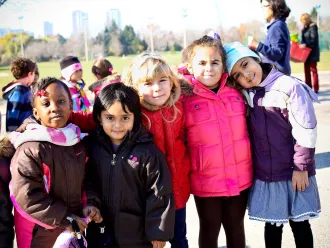 A diverse group of children stand shoulder to shoulder outside on a chilly but sunny day, smiling at the camera.