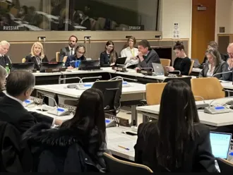 A group of delegates sit around a conference table with nameplates, engaged in discussion. The setting is formal, suggesting a diplomatic meeting.
