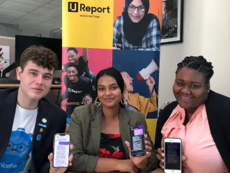 Three young people holding up cellphones in their hands and smiling. Behind them is a standee with a poster that shows the U-Report logo. 