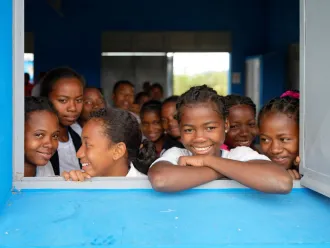 A group of more than ten smiling children look outdoors from a classroom window.