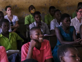 A classroom in Ghana with students sitting at desk. In the centre of the photo is a girl in a pink uniform smiling while looking beyond the camera. 