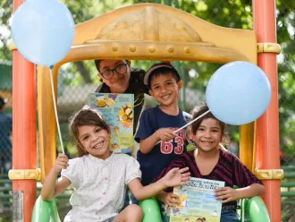 Four children stand together on a play structure and smile while holding balloons and reading materials.