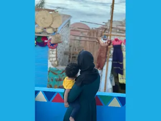 A woman looks out from a balcony, her back is towards the camera and she is holding a child in her arms. 