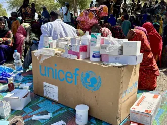 A UNICEF box covered with medical supplies sits on a colorful mat. In the background, people in vibrant clothing gather, conveying a sense of community.