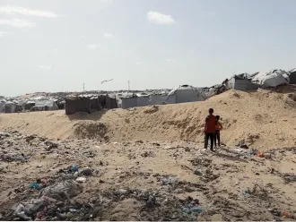 Two children stand in a sandy field with garbage around overlooking tents