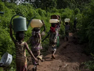 Children walk along a path in the forest, carrying buckets atop their heads, to gather water from a natural spring at Elevage IDP camp in Bambari, Central African Republic