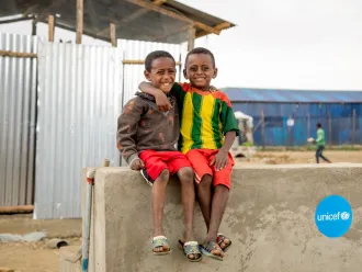 Two children sit on a concrete wall with their arms around each other’s shoulders and smiling at the camera.