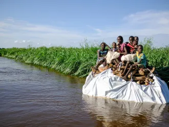 A group of children sit on top of a tarp of belongings on the sides of a flooded river bank.