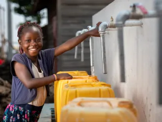 A girl smiles at the camera while filling a yellow water jug at an outdoor tap.