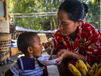 A woman in a red sweater feeds a young child with a spoon from a bowl; bananas and baskets are nearby, and trees are visible in the background.