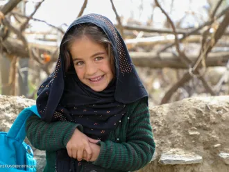 En Afghanistan, une jeune fille sourit à la caméra, la sangle bleue d’un sac à dos de l’UNICEF sur le bras.
