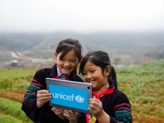 Two girls stand outdoors in a mountainous green field, looking at a tablet with a UNICEF logo on it. This photo was taken in Vietnam. 