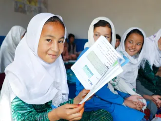 At a classroom in Afghanistan, a girl wearing a white headscarf holds open a book and smiles at the camera.