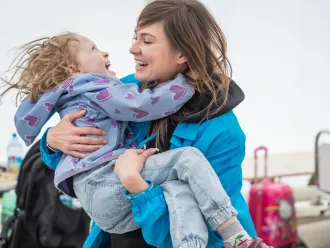 A woman wearing a UNICEF jakcet carries a child in her arms, both the woman and child are laughing. They are outdoors and both are wearing jackets.