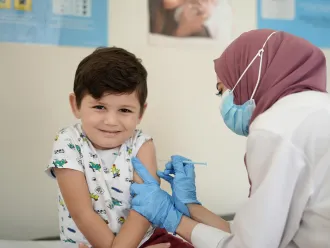 A child looks at the camera with a shy smile, a health care worker in a mask and medical gloves gives him a shot in the arm. The setting looks like a clinic. 