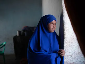 A girl wearing a blue headscarf look out of a window. A curtain is silhouetted in the foreground on the right side of the photo. 