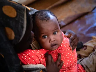 A child wrapped in a red polka dotted blanket looks up to the camera. he is held by his mother whose face is cast down.
