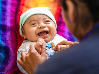 A woman plays with a baby lying on its back, she is holding his hands and the baby is smiling.