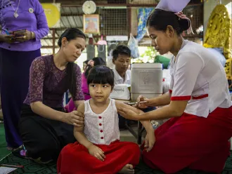 A girl sitting cross legged, looking very calm while getting an injection by a nurse, another woman is next to the child, looking at her. 