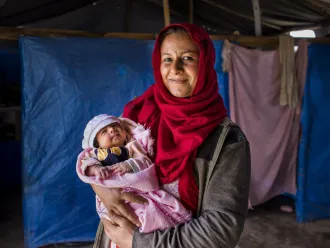A mother in front of a tent holding a newborn in pink blanket. 
