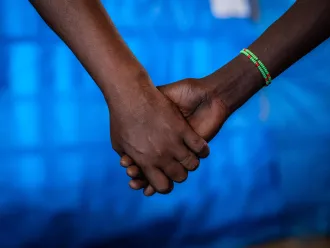 Hands being held, the hand on the left is that of a man and the one on the right of a girl. The girl is wearing a green beaded bracelet. The photo is cropped till the forearms, the background is that of a blue tarp.