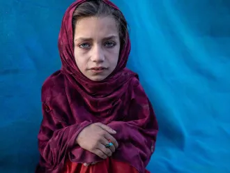 A girl wearing a magenta headscarf while sitting, looking at the camera, with her hands folded on her knees. There is a vivid blue tent behind her