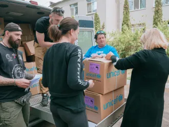 A van with the back door open and a several boxes with the words UNICEF written on them. Two women stand in front of the boxes looking at them. One man is unloading the boxes and two other men are standing to the side.