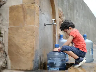 a boy squats in front of a water tap which is filling up a gallon bottle with water. 