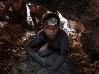 A young person wearing a headlamp crawls through a narrow, rocky cave. Behind them, others follow. The atmosphere is tense and focused.