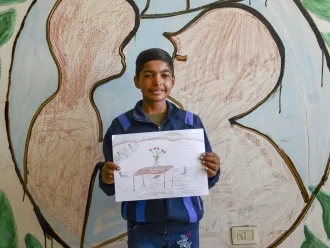 A boy holds a drawing up. It is made on paper and with colour pencils, showing a table with a vase and flowers on it. The boy is looking at the camera smiling. 